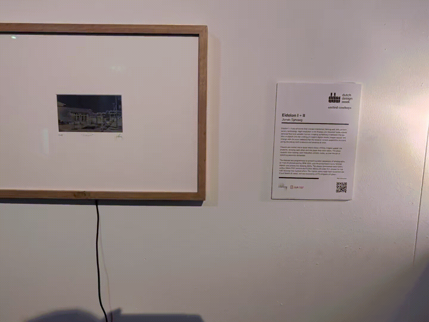 Framed artwork with small blue-toned image displayed on white wall next to exhibition label from Dutch Design Week, connected by black cable