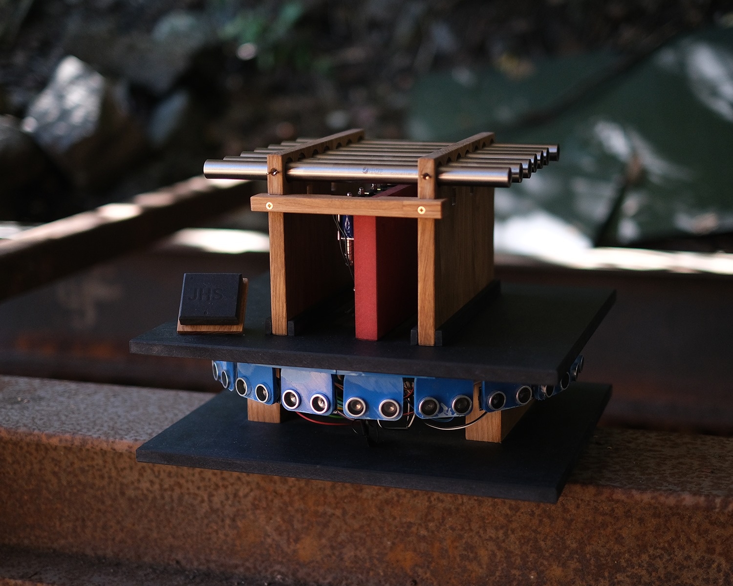 Wooden and blue mixed-media sculptural model with geometric structures and metal tubes, positioned on dark platform outdoors in natural lighting