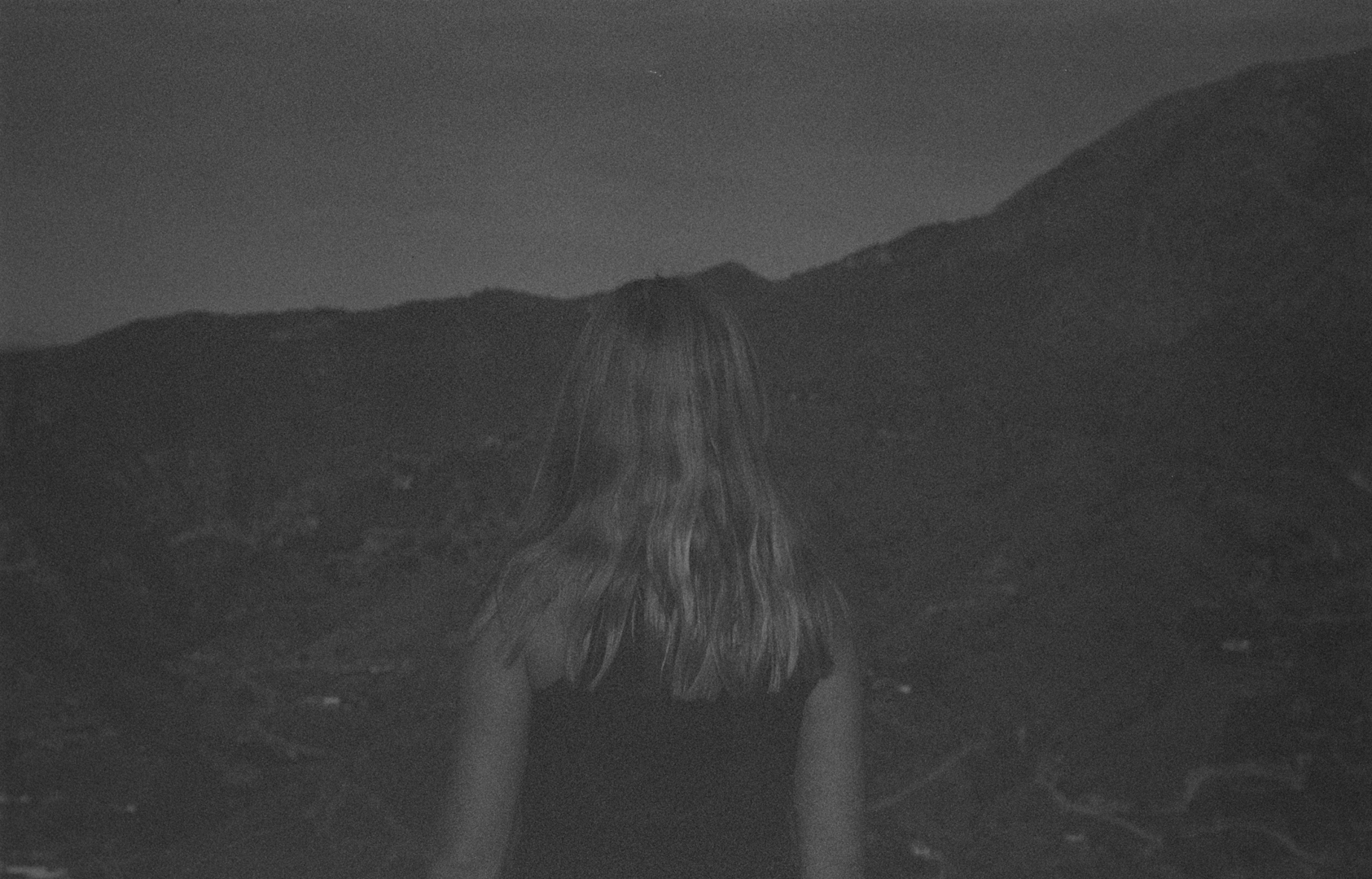 Black and white photo showing back of person with shoulder-length hair facing mountainous landscape in moody, contemplative composition