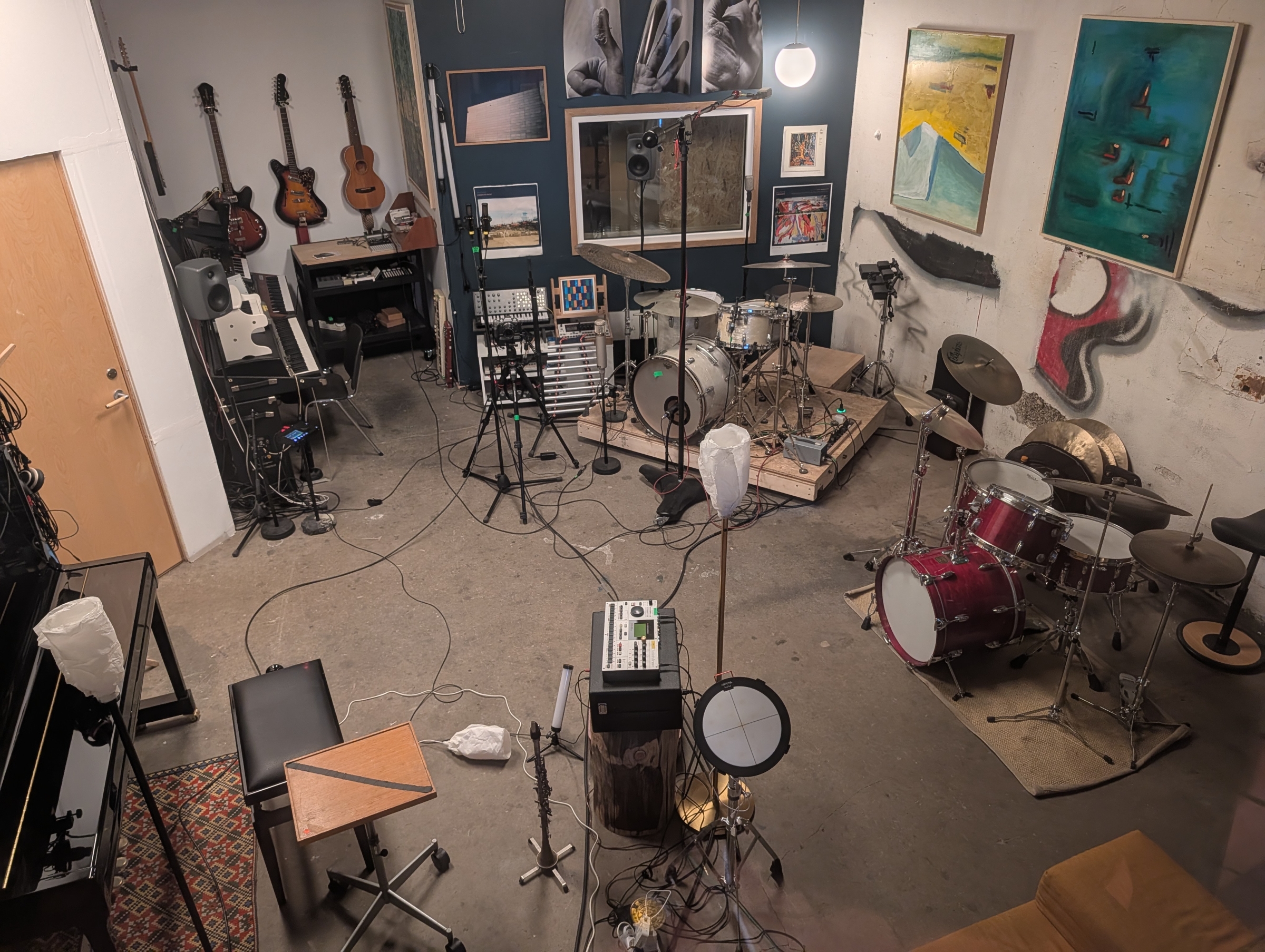 Music studio with drum kits, guitars on wall, recording equipment, microphones, artwork, and colorful paintings in cozy rehearsal space