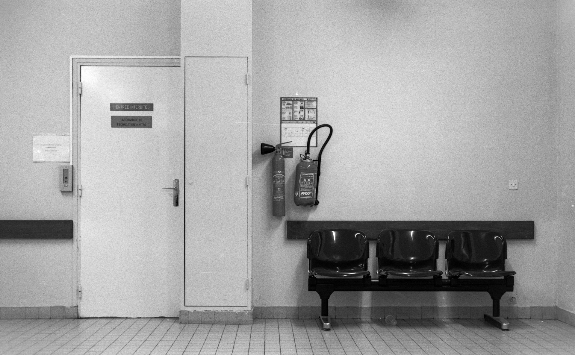 Black-and-white hospital corridor with "Entrée Interdite" door, fire extinguishers on wall, and three empty bench chairs on tiled floor.