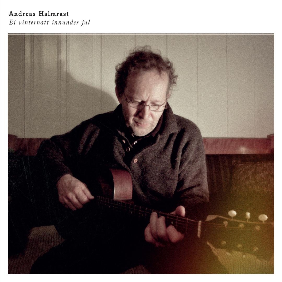 Elderly man with glasses and curly gray hair playing acoustic guitar, wearing dark sweater, seated against white wooden wall in warm indoor lighting