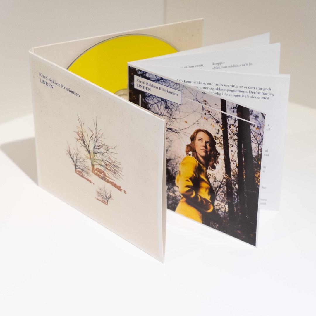 Album artwork for "Linden" by Kira Bakken Kristiansen showing white gatefold packaging with yellow vinyl, bare trees illustration, and woman in yellow coat photograph