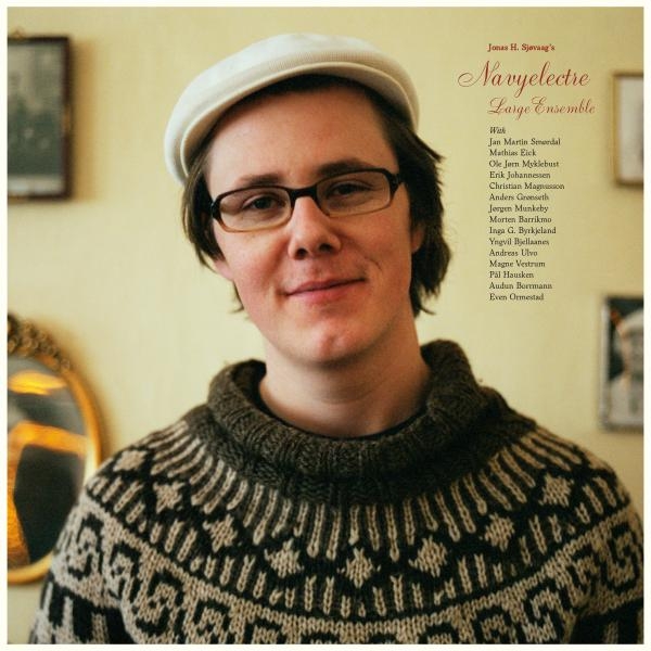 Person in patterned sweater and white cap smiling at camera, warm indoor setting with yellow walls and framed pictures, album cover layout