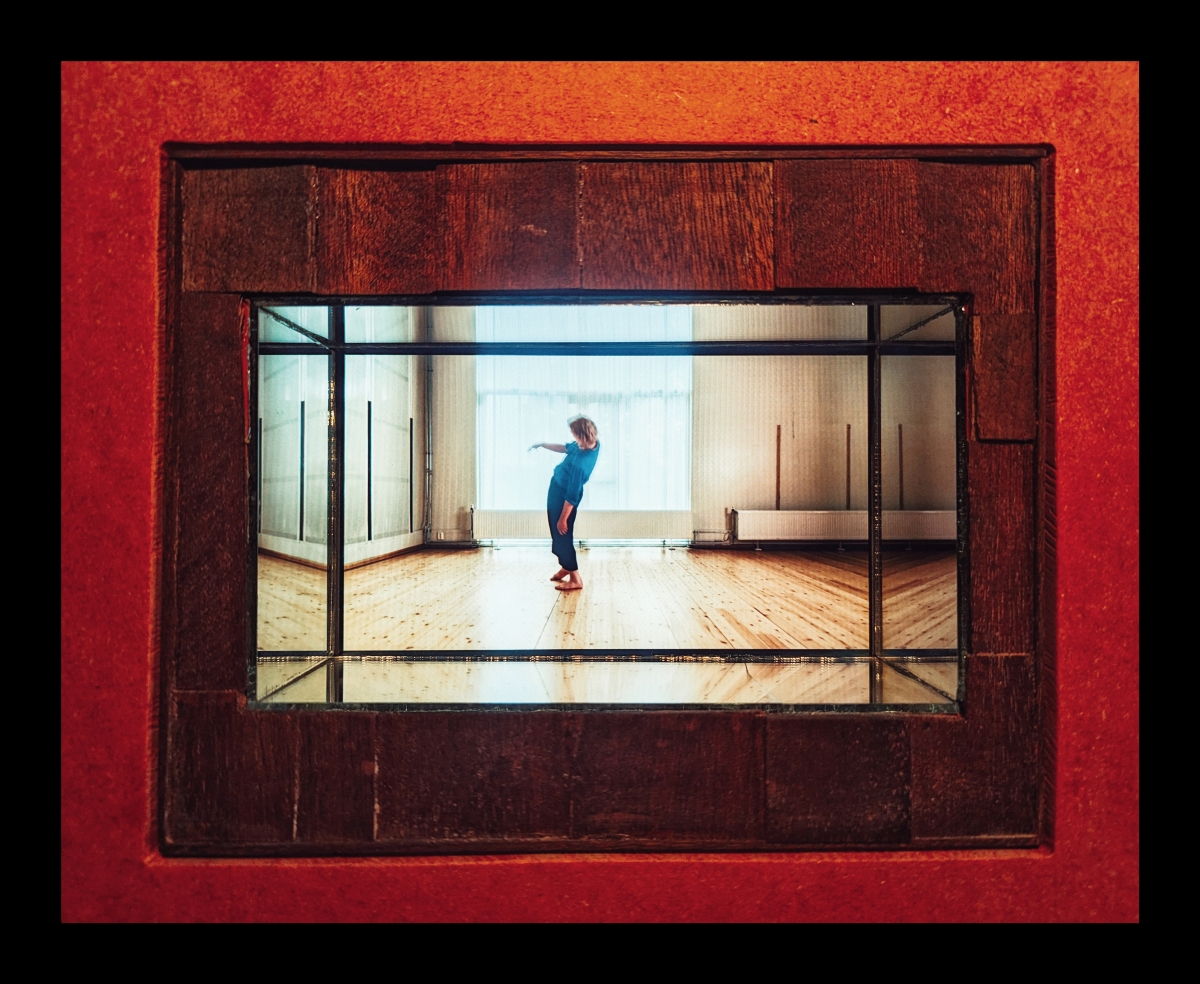 Red-framed vintage viewfinder showing person in blue shirt dancing in bright wooden studio space with warm lighting and windows