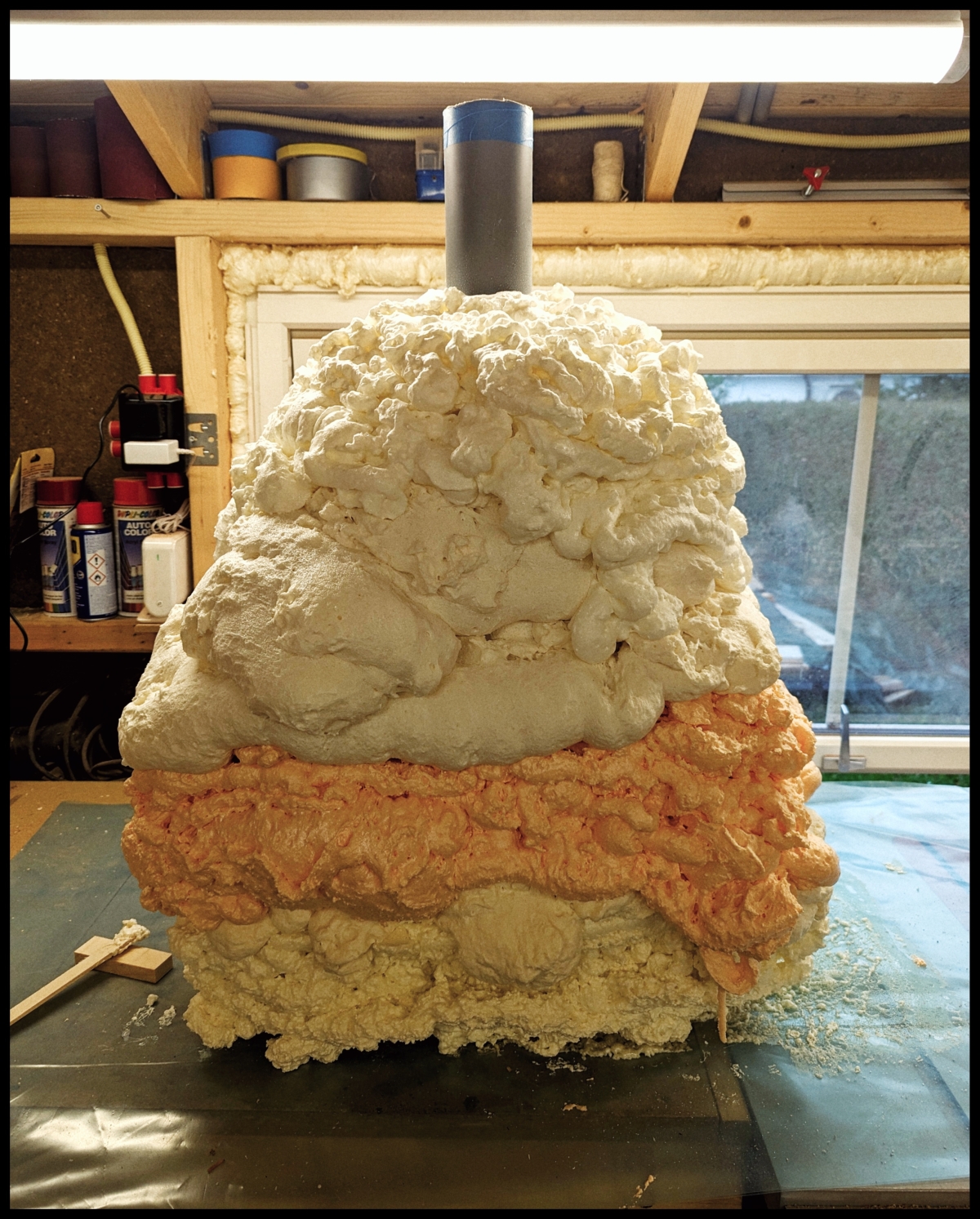 Workshop sculpture showing layered clay structure with white top section, orange middle layer, and beige bottom, with pipe extending upward in wooden studio space.