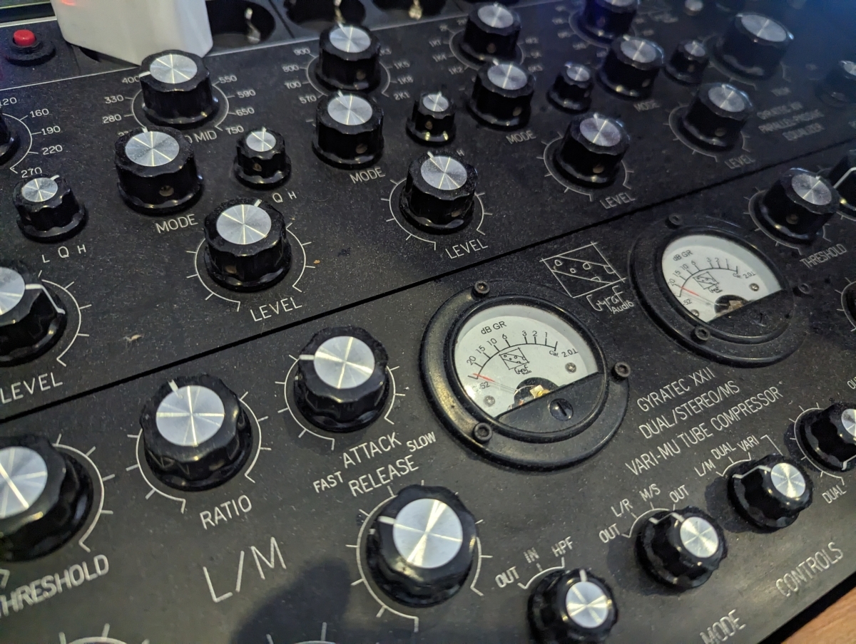 Black audio mixing console with silver knobs, analog VU meters, and control labels including "ATTACK", "RELEASE", "RATIO" and "THRESHOLD