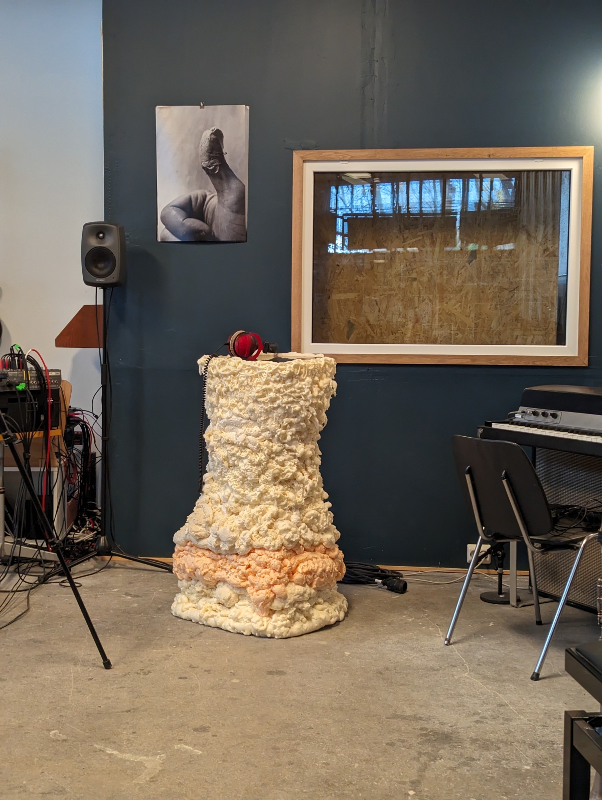Art studio with textured cream sculpture, monitor speaker, framed artwork on dark wall, and music equipment in industrial space.