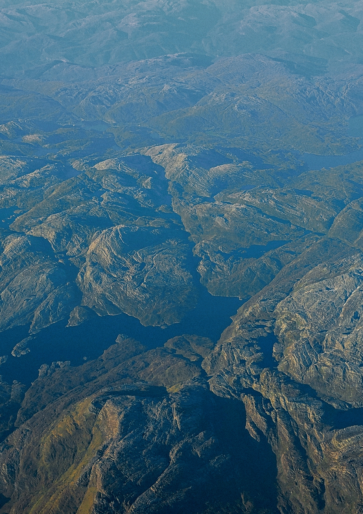 Aerial view of rugged mountainous terrain with deep valleys and ridges in muted blue-gray tones, creating a textured landscape pattern.