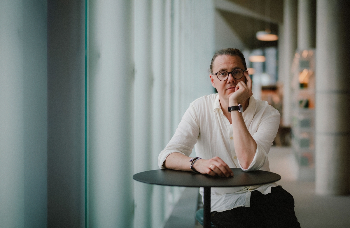Person with glasses wearing white shirt sits thoughtfully at dark round table, hand on chin, in modern space with large windows and soft lighting