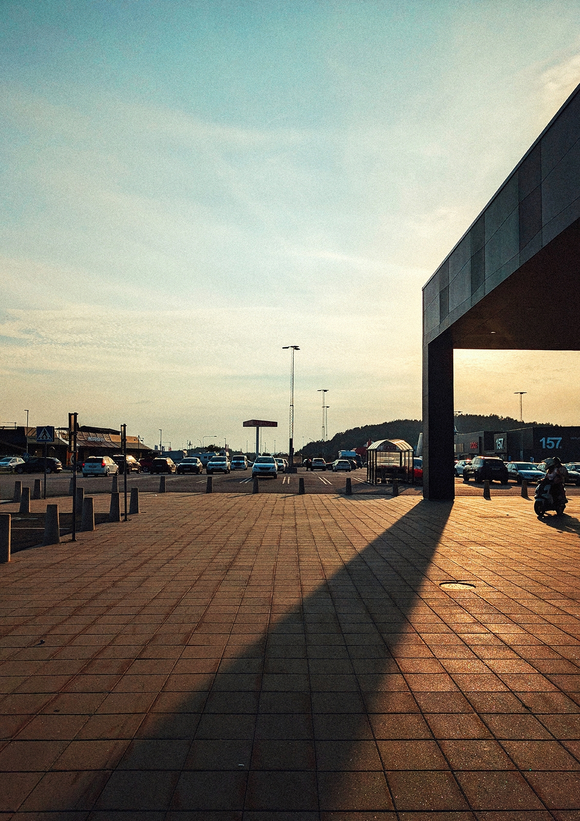 Golden hour at Swedish transportation hub with modern building casting long shadows across brick plaza, cars and buses visible under dramatic sky.