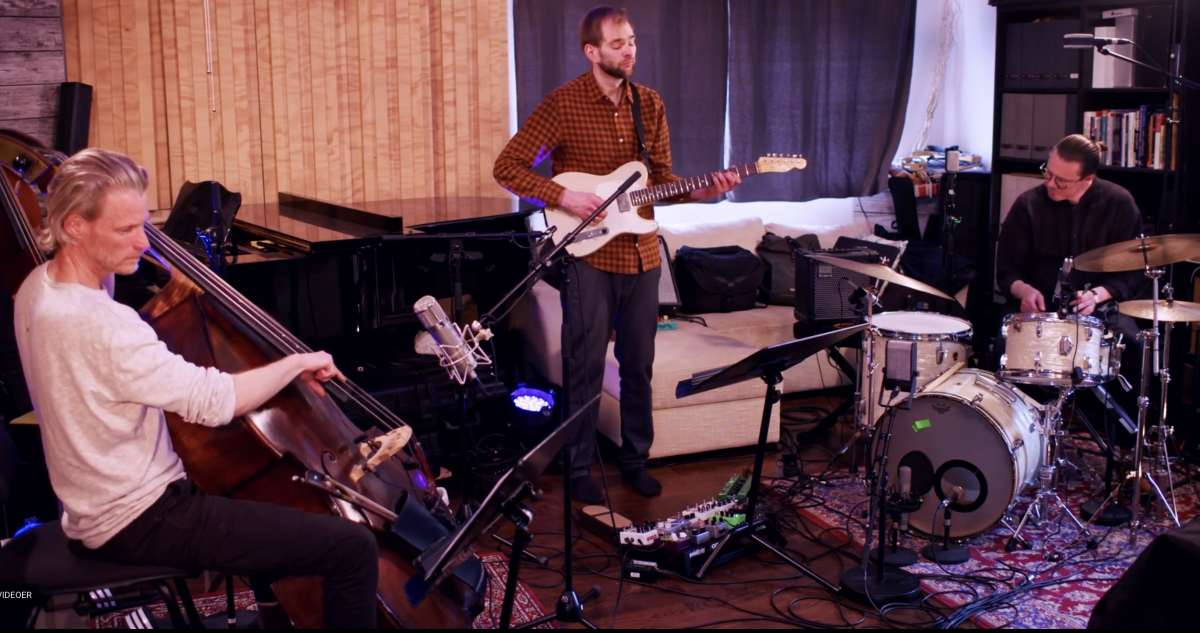 Three musicians performing in a cozy studio: blonde bassist in white sweater, guitarist in plaid shirt, and drummer in black, with warm lighting and colorful rug.