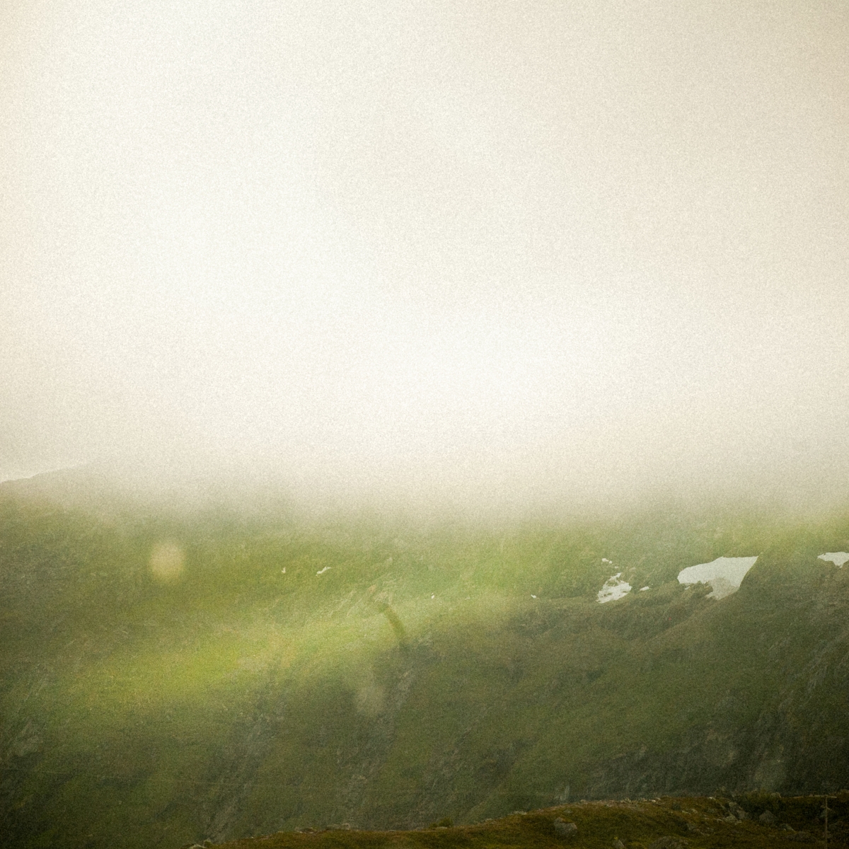 Misty green mountainous landscape in Norway with patches of snow visible through heavy fog, creating a moody atmospheric scene