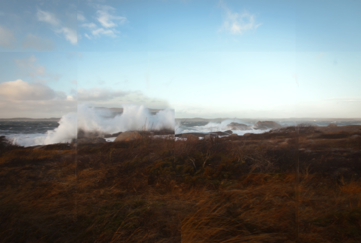 Dramatic coastal scene with large waves crashing against rocky shore creating white spray, brown vegetation in foreground under cloudy blue sky