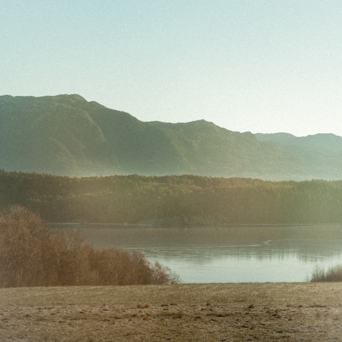 Misty mountain landscape with layered peaks in muted greens rising above a calm lake, surrounded by brown vegetation under pale sky
