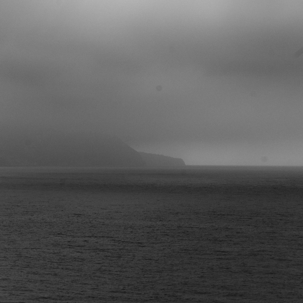 Moody black and white seascape showing choppy dark waters under heavy gray clouds with distant mountainous coastline shrouded in mist.