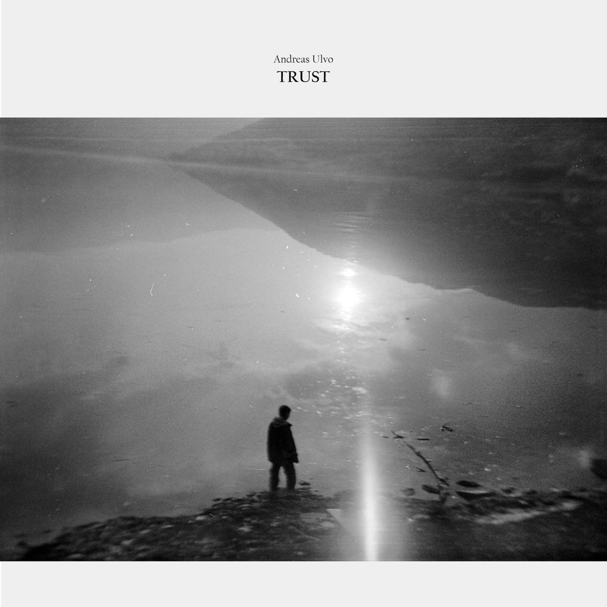Black and white photograph showing silhouette of person standing on rocky shore with dramatic cloudy sky and sunlight reflecting on water below