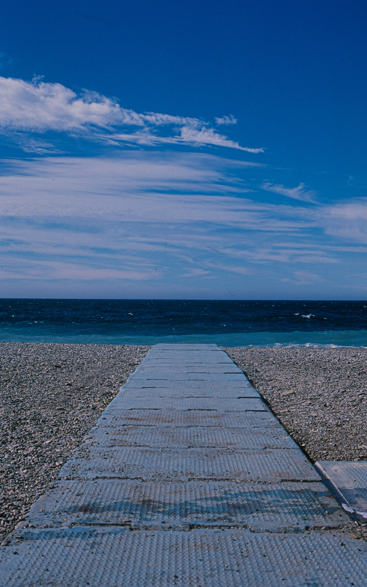 Wooden boardwalk leads across pebble beach to deep blue ocean under dramatic sky with wispy white clouds stretching across bright blue expanse