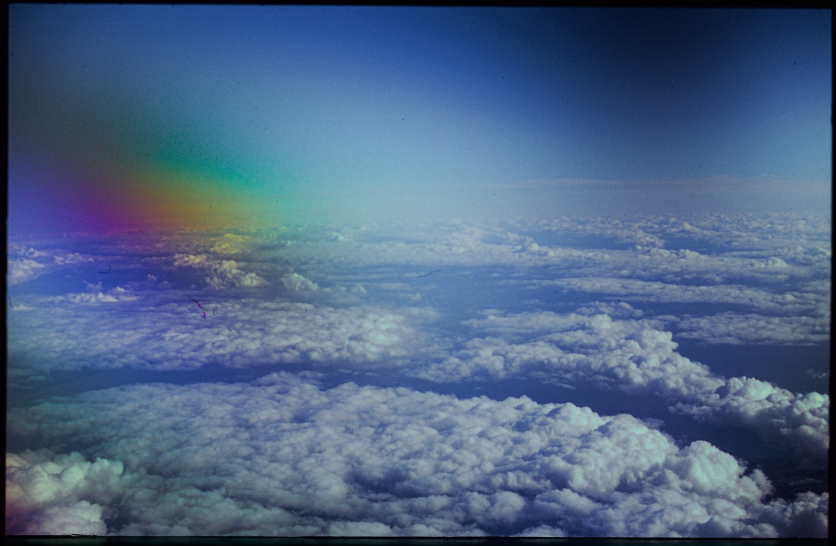 Aerial view of fluffy white clouds stretching to horizon under blue sky with rainbow light prism creating colorful gradient in upper left corner