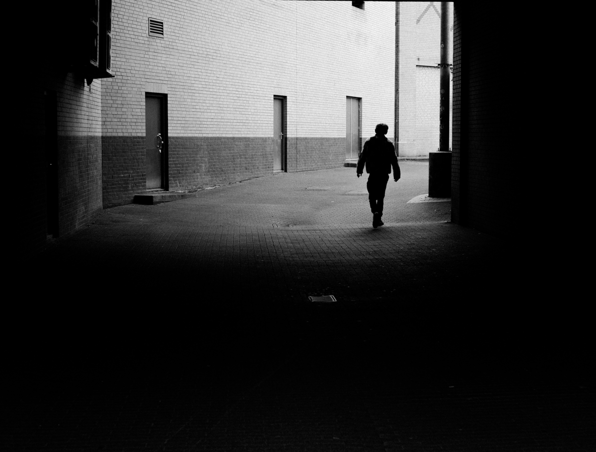 Silhouetted figure walks through shadowy brick alleyway toward bright sunlit opening, creating dramatic black and white contrast and moody urban atmosphere.