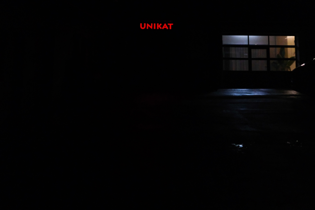 Dark nighttime scene with illuminated building windows and "UNIKAT" text in red, creating moody atmospheric lighting against black background.