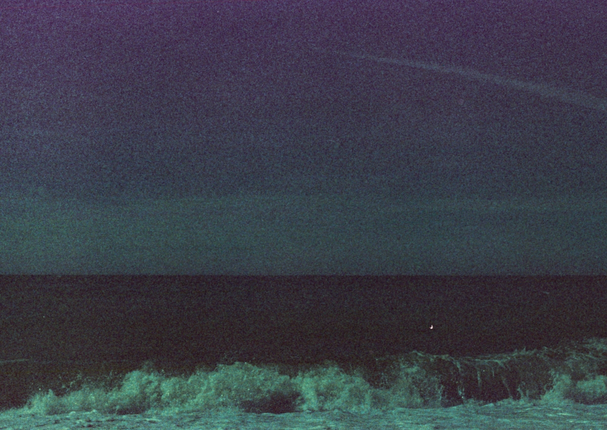 Grainy expired-film seascape at dusk, teal-toned sky over dark ocean, pale foam waves breaking along the shoreline.