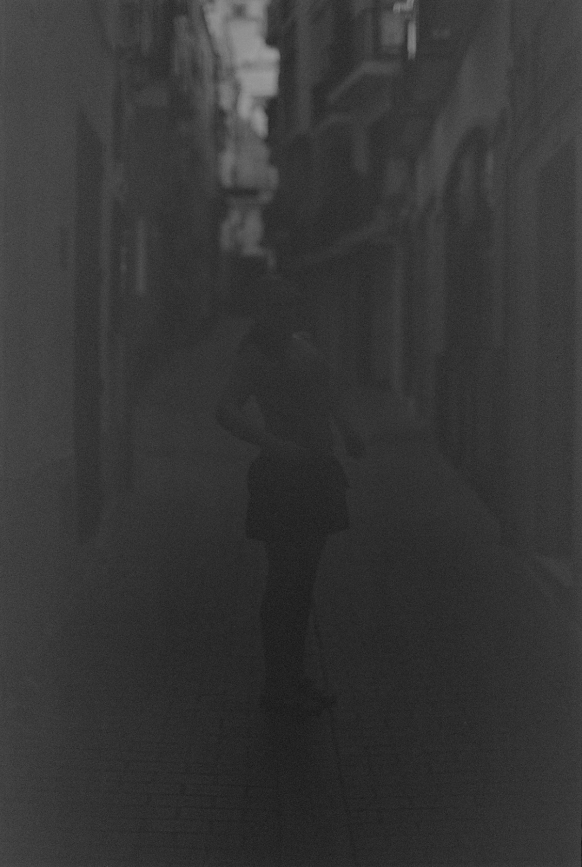 Dark, moody black and white street scene with narrow alleyway between tall buildings, creating dramatic shadows and atmospheric urban composition.