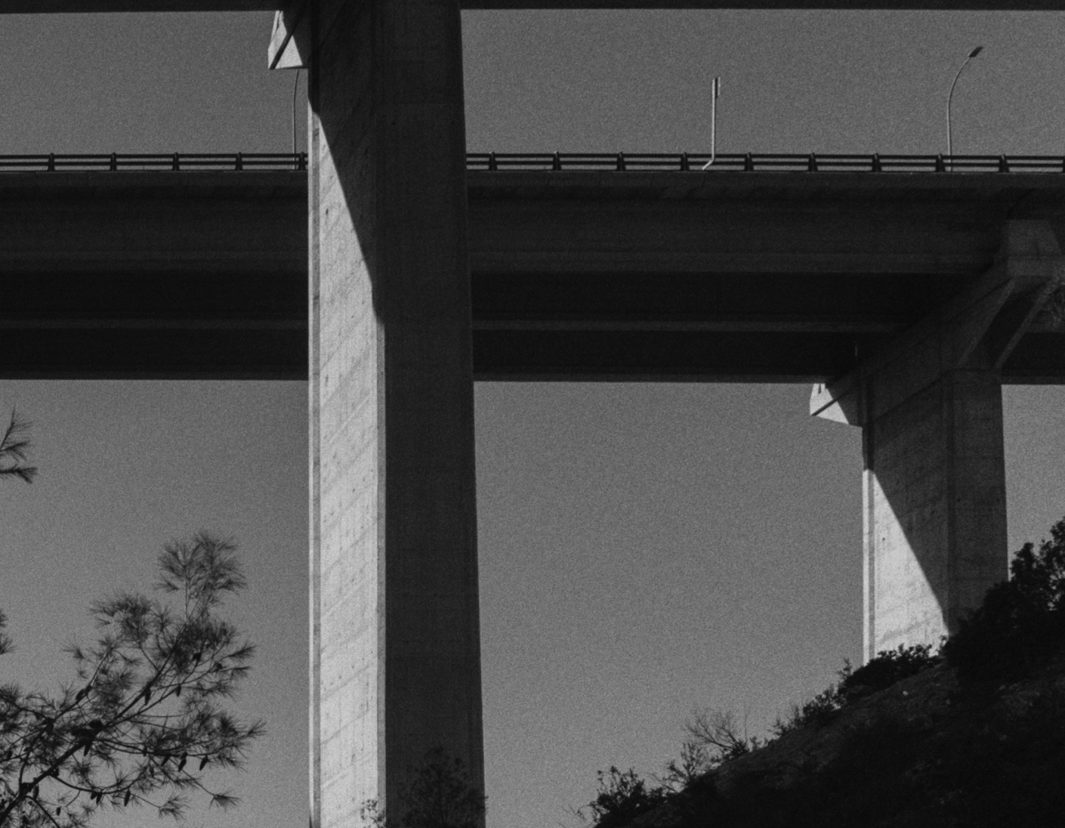 Black and white architectural photograph showing concrete bridge pillars creating geometric shadows against gray sky, with vegetation silhouettes below