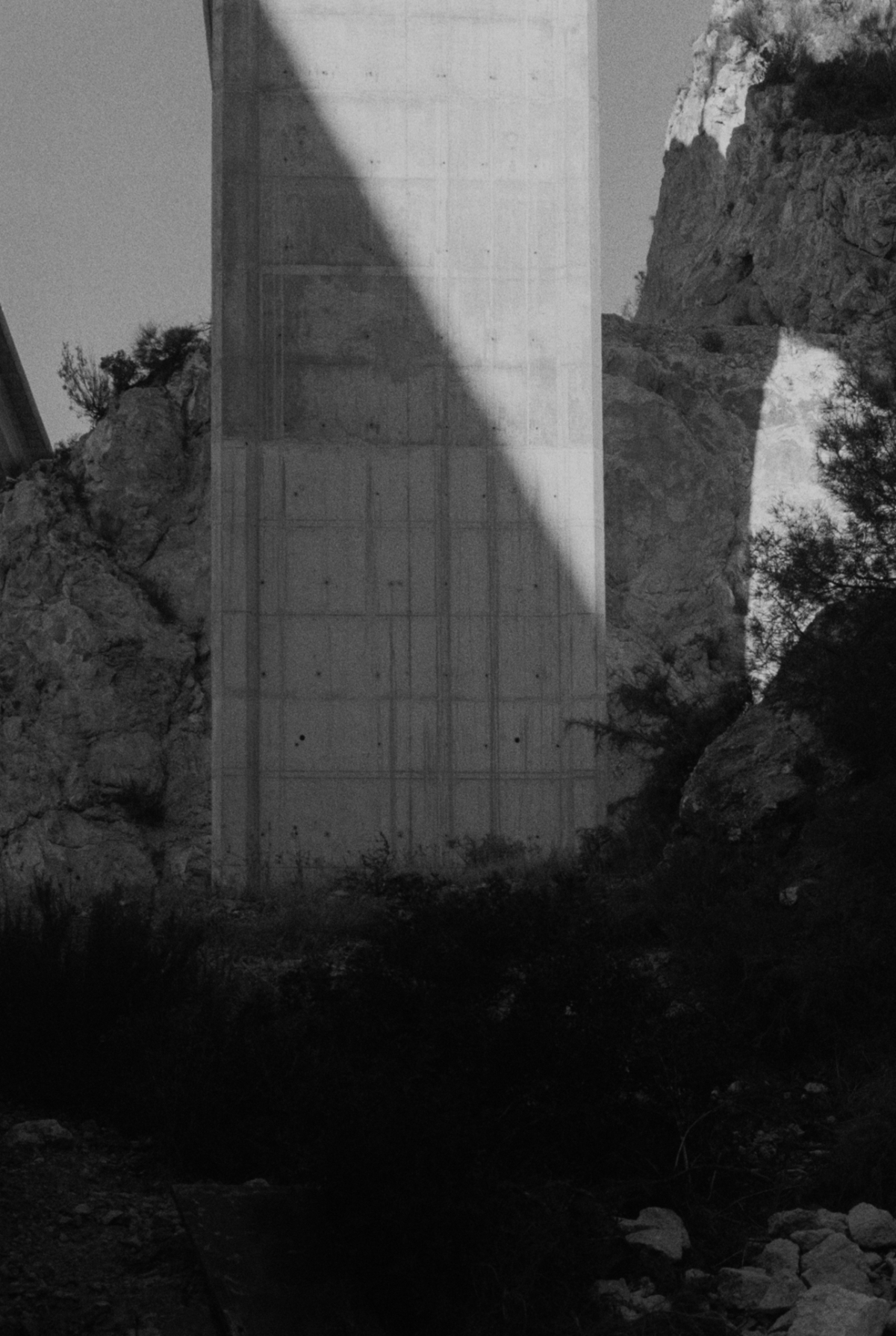 Black and white photograph showing a concrete dam structure between rocky canyon walls, with dramatic shadows and natural vegetation in the foreground.