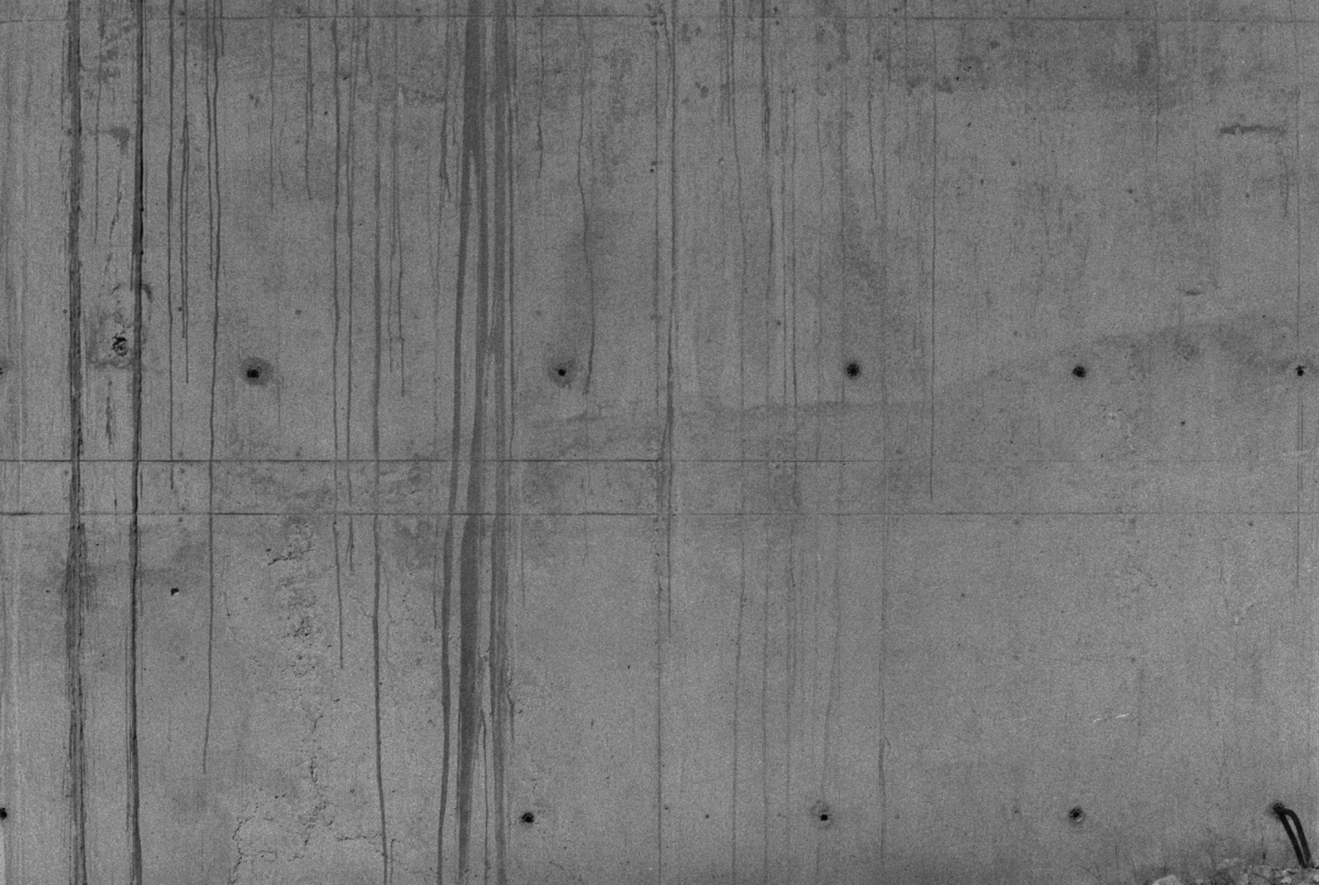 Raw concrete wall with vertical panels, dark staining streaks, and circular tie holes creating geometric industrial pattern in grayscale