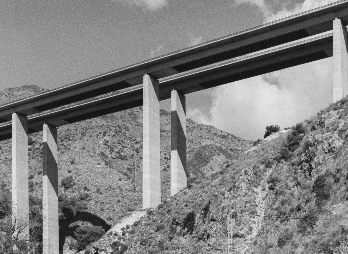 Black and white photograph of a concrete highway bridge with tall pillars spanning across a mountainous valley with vegetation-covered hills.