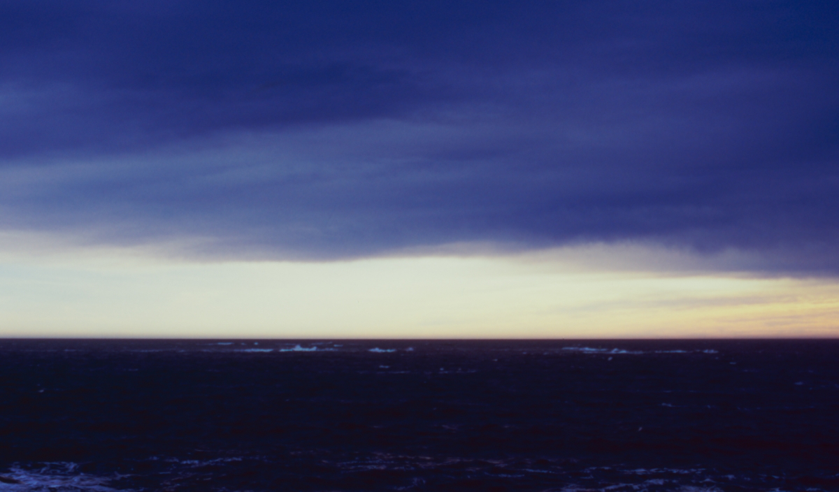 Dark ocean under dramatic purple and blue storm clouds with pale yellow horizon glow, creating moody seascape at dusk or dawn