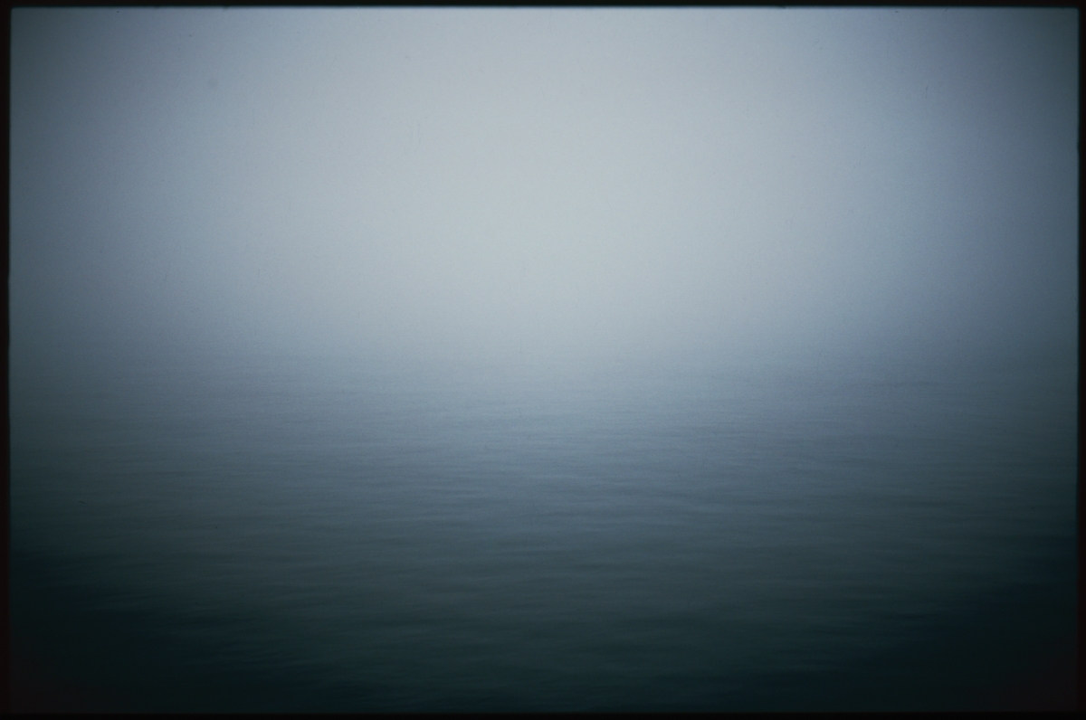 Misty seascape with calm dark waters meeting foggy gray sky, creating moody atmospheric scene with muted blue-green tones