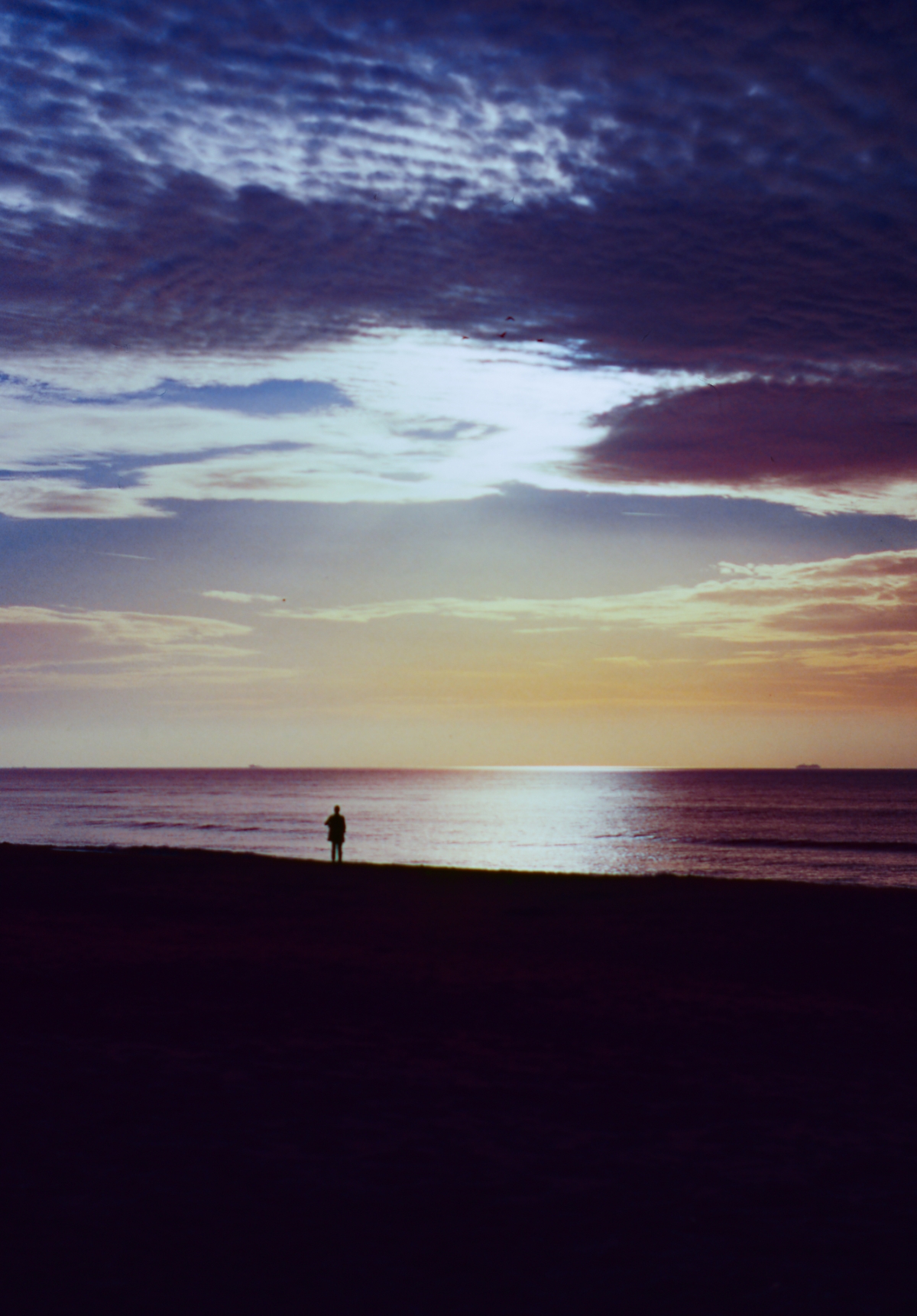 Solitary figure silhouetted on beach at sunset with dramatic purple and orange clouded sky reflected on calm ocean water