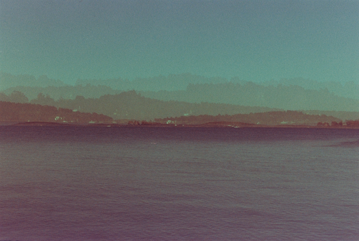Faded coastal landscape shot on expired film, with muted teal sky, hazy tree-lined hills, distant shoreline lights, and calm rippling water below.