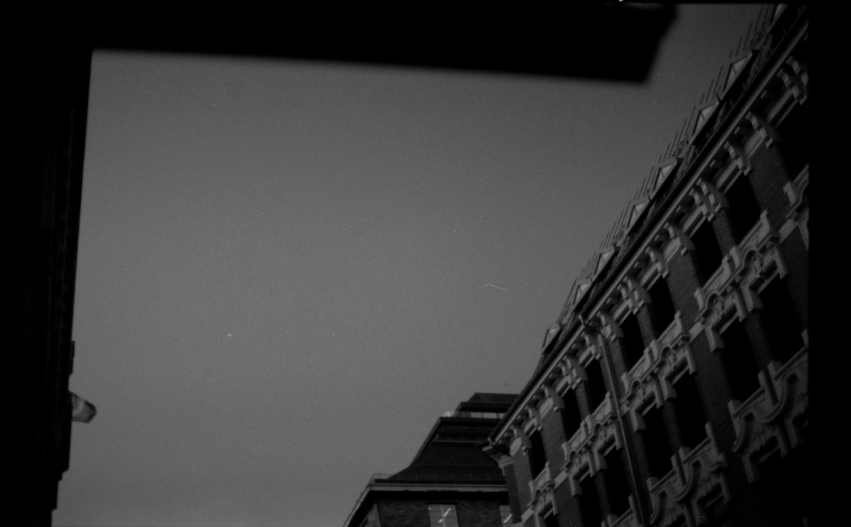 Black and white upward view of tall brick buildings creating dramatic angular perspective against overcast sky from street level