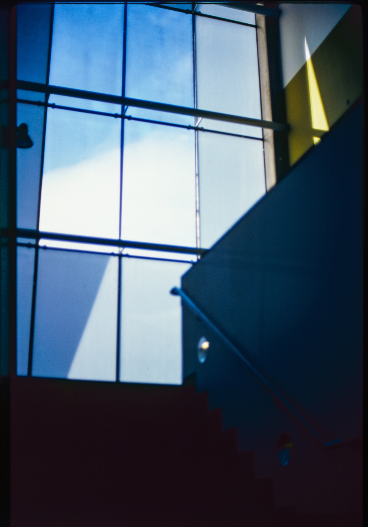 Blue-tinted interior view showing stairs ascending toward large gridded windows with bright sky visible, creating dramatic shadows and geometric light patterns.