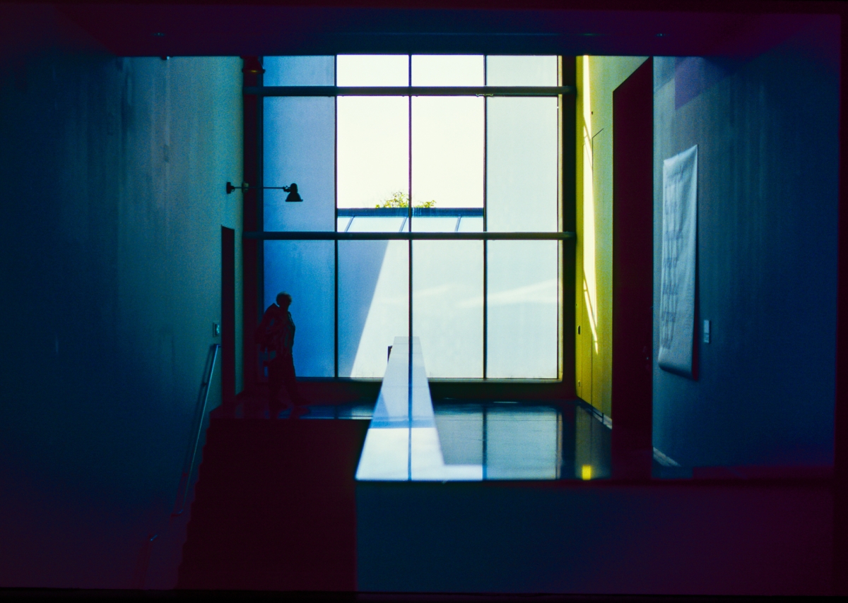 Modern interior with dramatic blue lighting, silhouetted figure by large grid windows, yellow accent wall, creating moody architectural composition