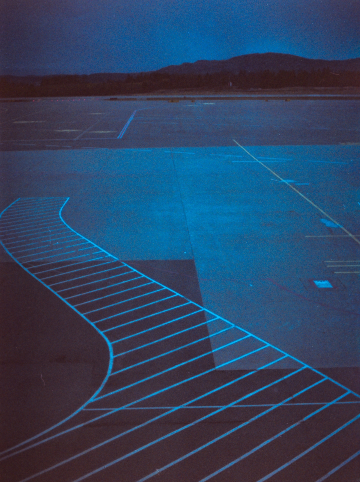 Nighttime airport tarmac with glowing blue chevron markings curving across pavement, distant runway lights, and dark silhouetted hills under twilight sky.