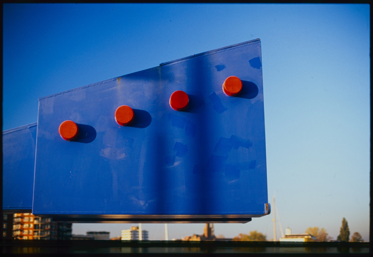 Blue billboard with four red circular lights against clear sky, urban buildings visible in background, film photography aesthetic