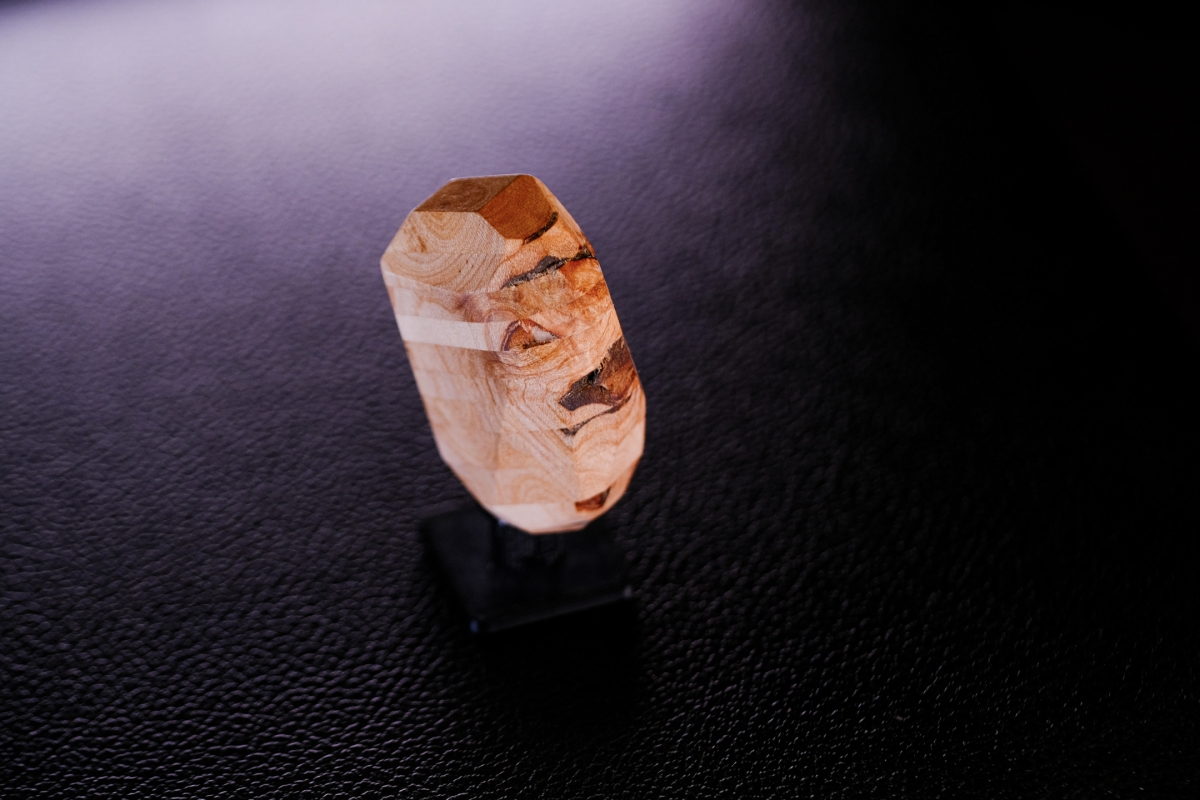 Polished wooden sculpture with natural grain patterns and geometric faceted top, casting shadow on dark textured surface under moody lighting