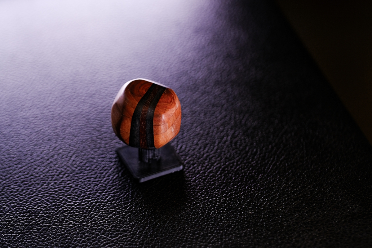 Polished wooden sculpture with dark grain stripes sits on black textured surface under dramatic purple-tinted lighting, creating moody shadows