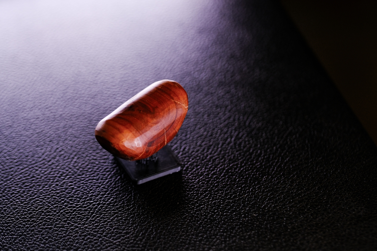 Polished red-orange wooden sculpture with natural grain patterns sits on black textured surface under dramatic lighting creating shadows and highlights