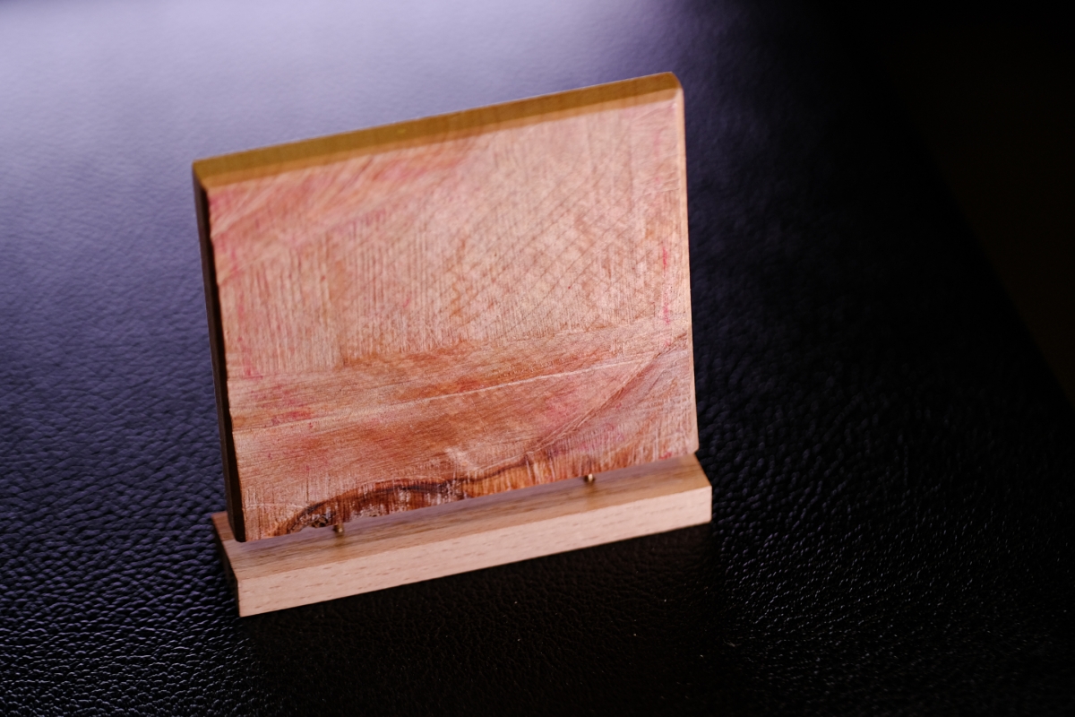 Wooden sculpture with curved form displaying natural wood grain patterns in warm tones, mounted on base against dark textured surface