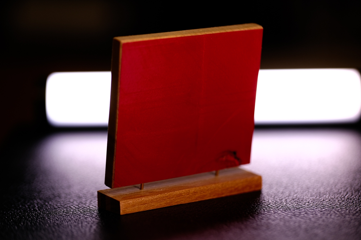 Red wooden block standing upright in natural wooden base on dark textured surface, backlit by soft white light creating dramatic shadows