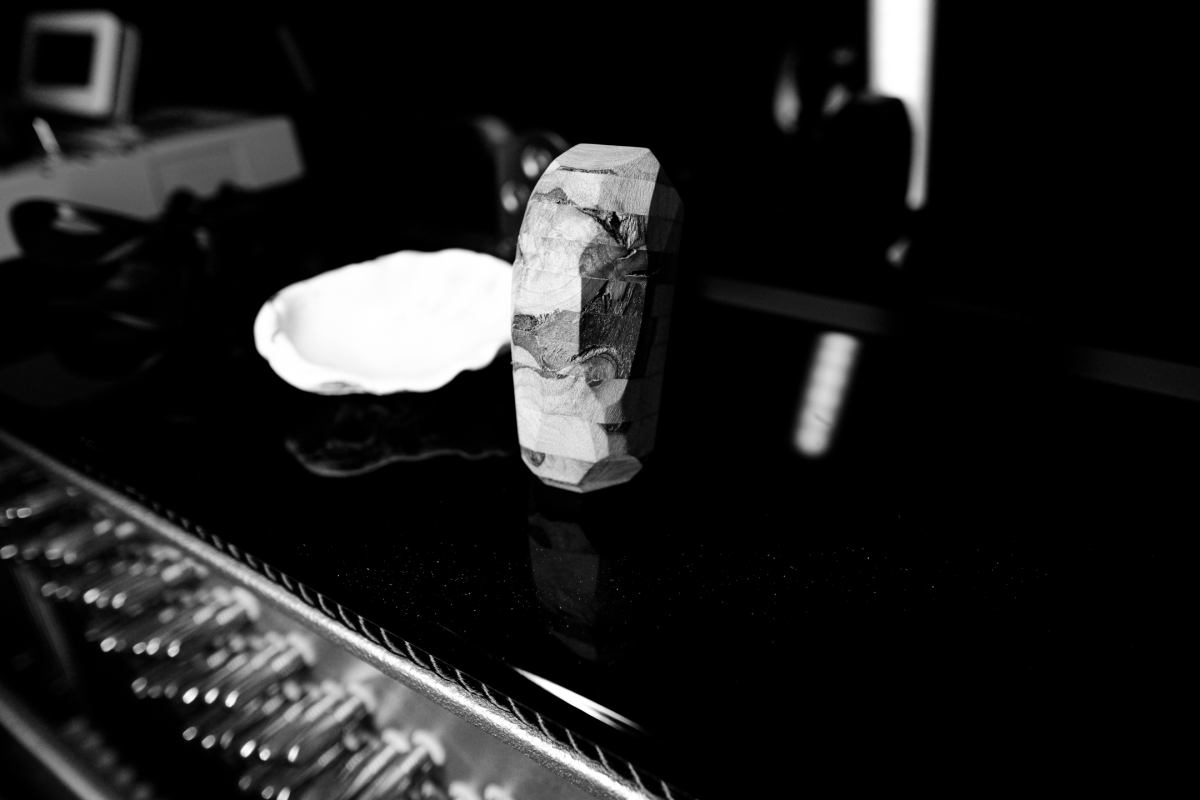 Black and white photo showing a rough-textured cylindrical stone sculpture on dark reflective surface with blurred equipment in background