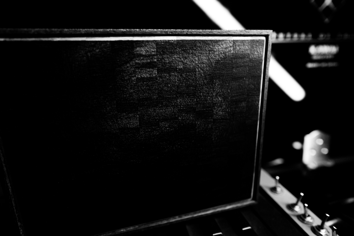 Black and white close-up of textured dark rectangular sculpture surface with blurred mechanical elements and white details in background