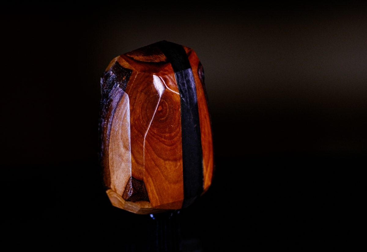 Polished wooden sculpture with rich orange-brown grain and dark purple-black accents, dramatically lit against black background