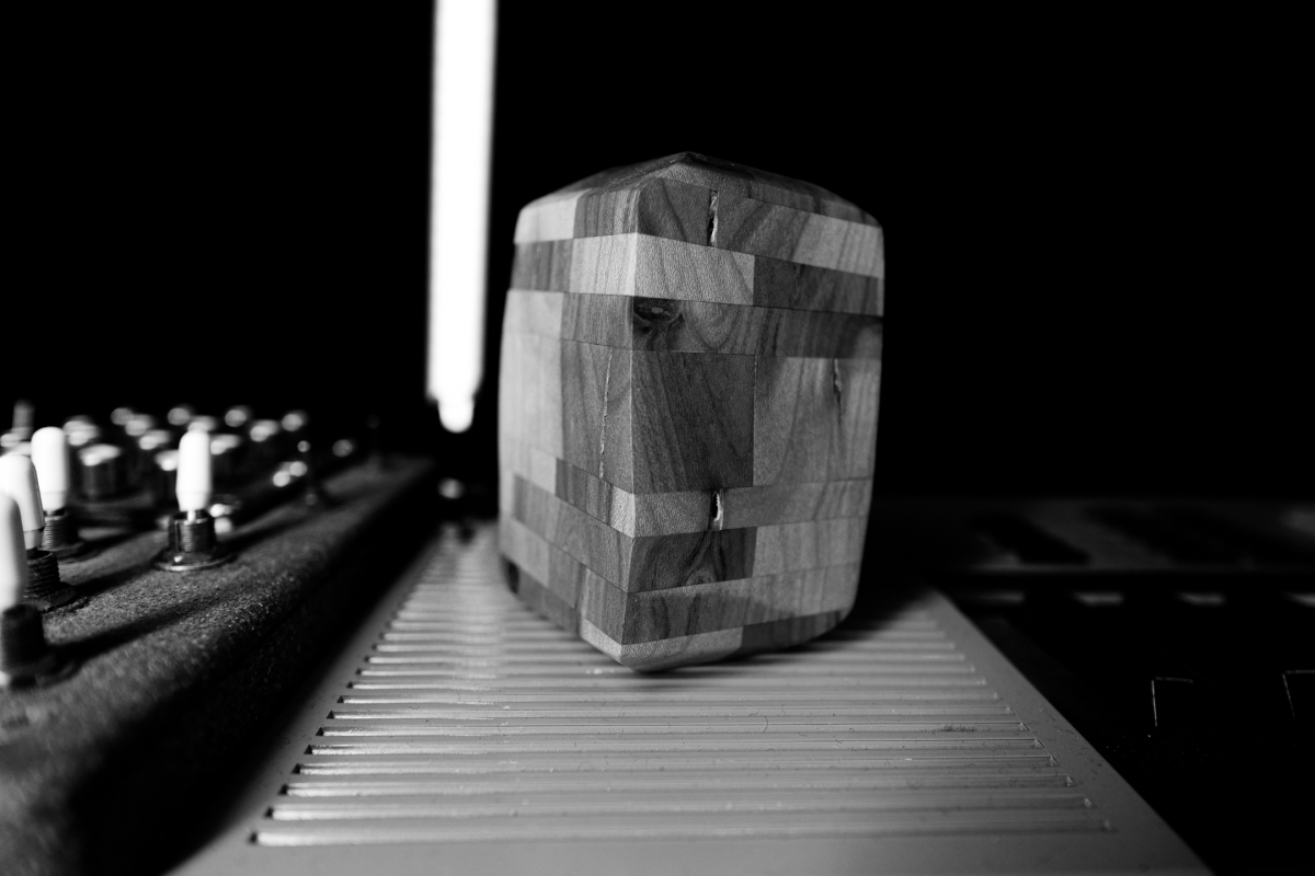 Black and white photo showing a geometric wooden block sculpture sitting on piano keys, with mixing board controls visible in background