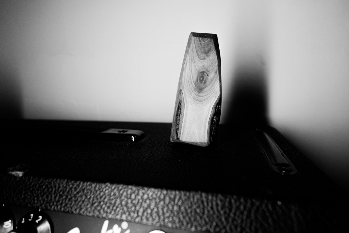 Black and white photograph showing a tall wooden sculptural object with visible wood grain standing upright on a dark textured surface against a blurred background.
